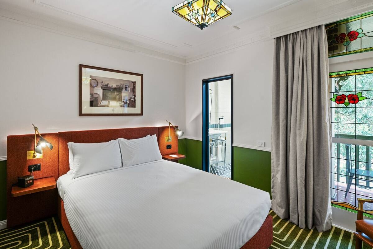 Una habitación de hotel con una cama, lámparas de noche, un cuadro enmarcado, una ventana con cortinas y una pared verde.