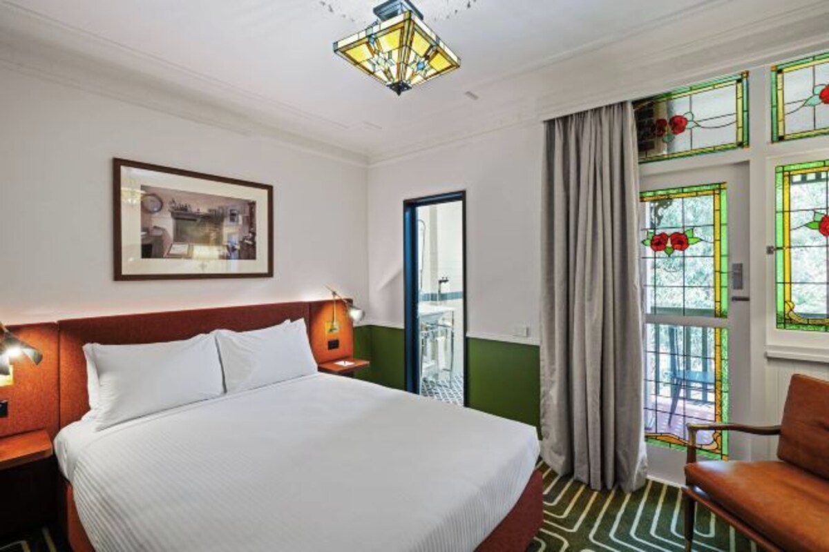 Una habitación de hotel con una cama, una mesita de noche, un cuadro enmarcado en la pared, un candelabro, una ventana con cortinas y una silla.