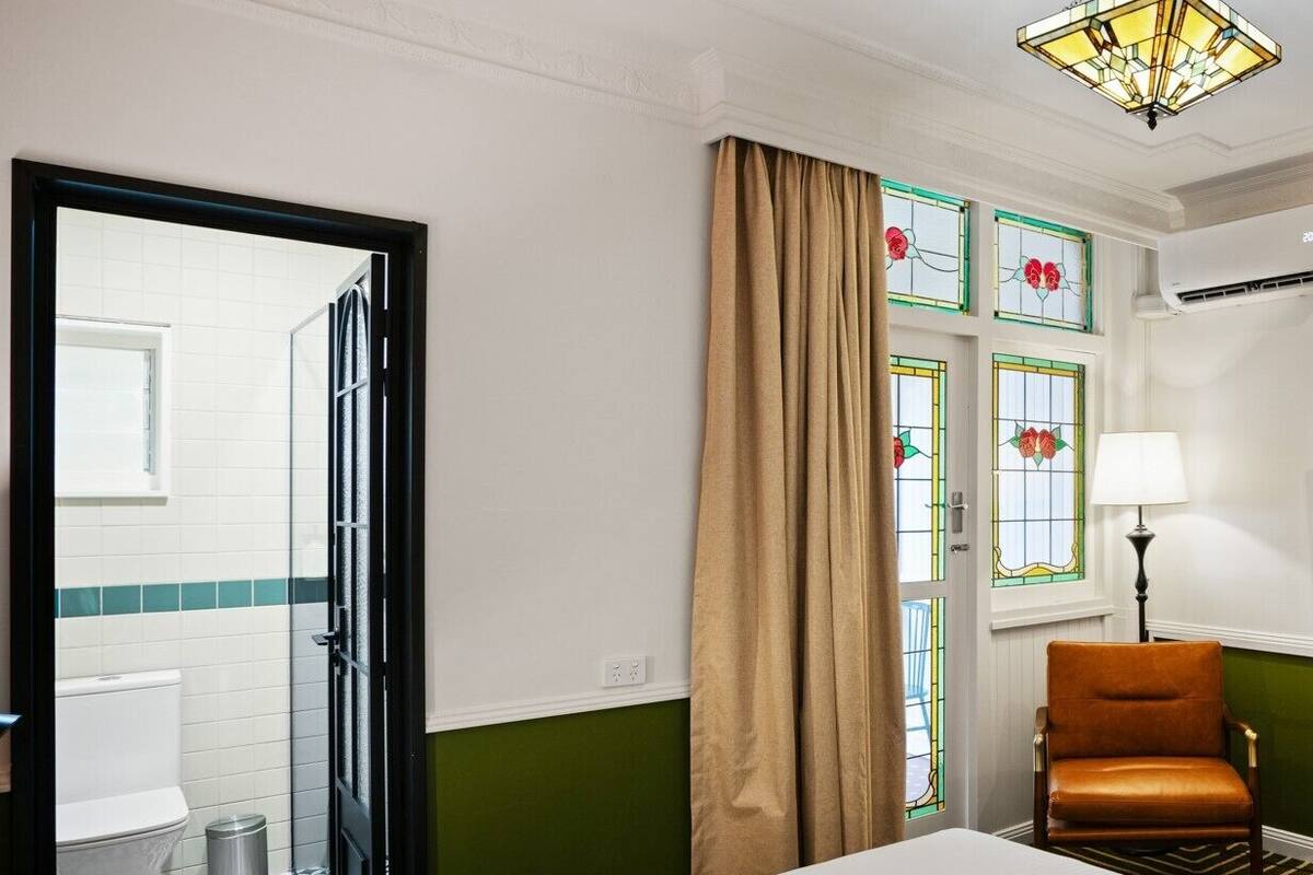 Habitación de hotel con cama, una silla, ventana con cortinas, lámpara y baño visible a través de una puerta abierta.