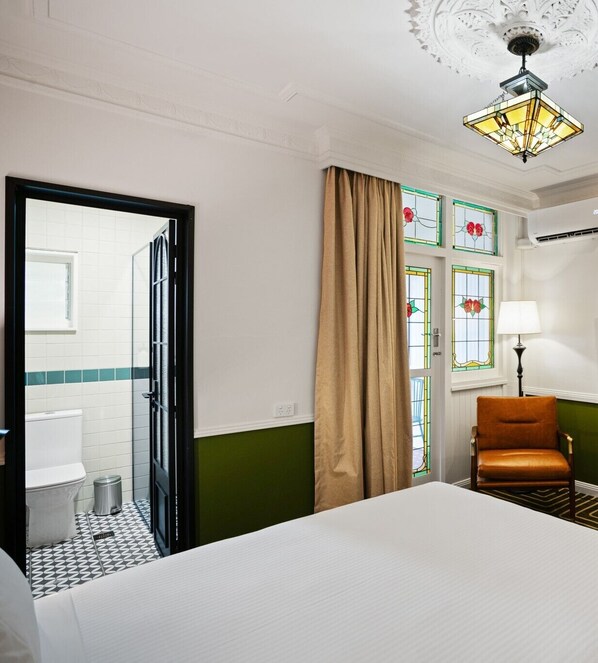 Habitación de hotel con cama, una silla, ventana con cortinas, lámpara y baño visible a través de una puerta abierta.