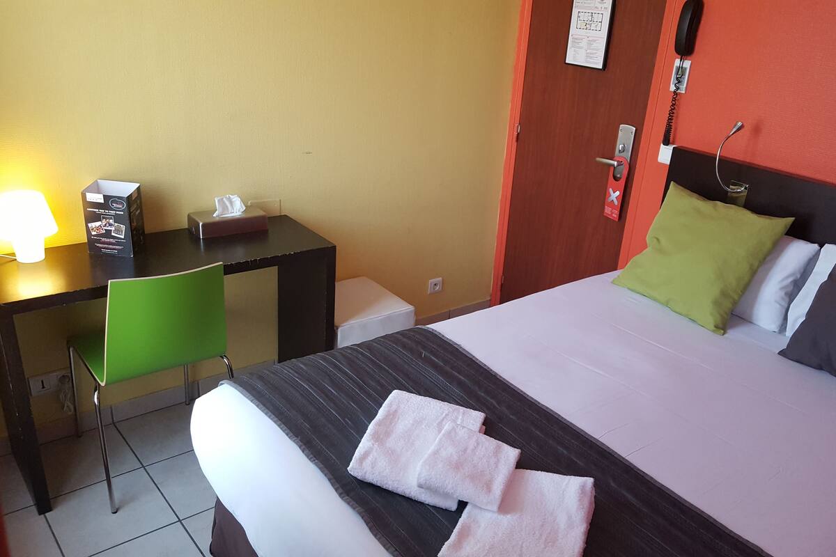 A hotel room with a bed, a desk, a chair, a lamp, and a telephone.