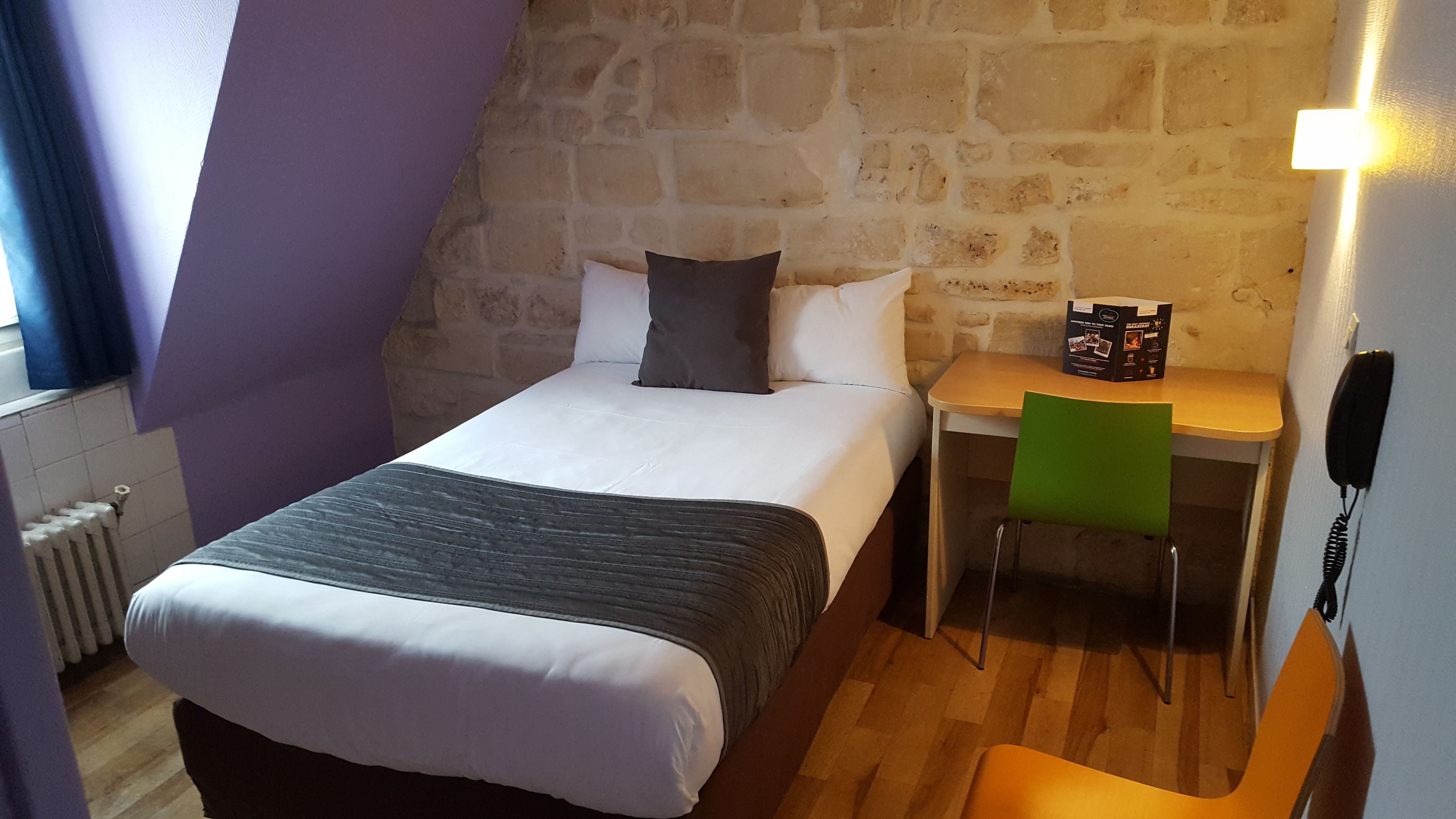 Ein Schlafzimmer mit Bett, Schreibtisch und Stuhl. Eine Steinwand befindet sich hinter dem Bett, und ein Fenster mit Vorhängen ist vorhanden.