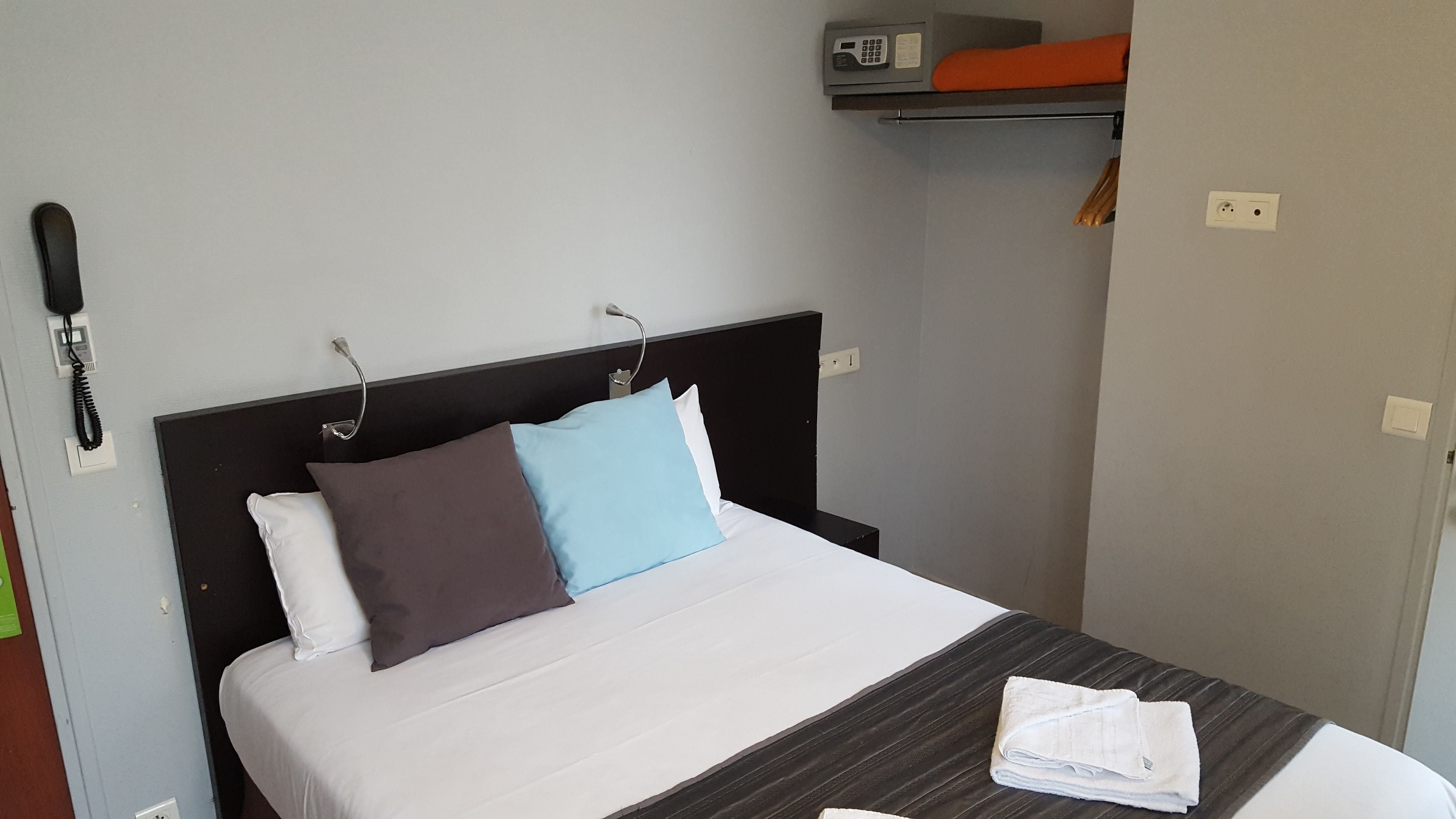 A hotel room with a bed, pillows, a folded towel, a telephone, and a shelf with a blanket.