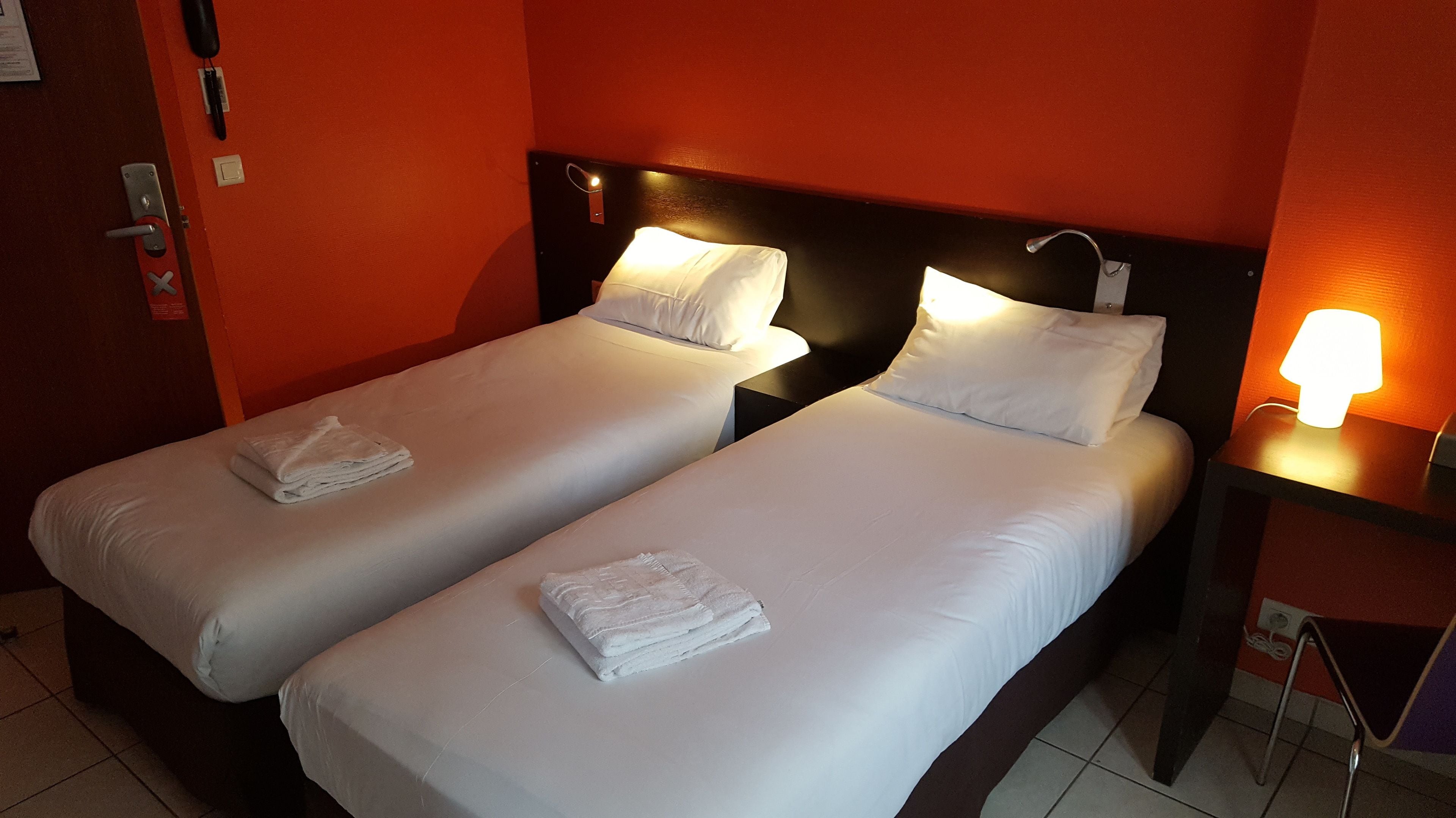 Two single beds with white linens and pillows, a nightstand with a lamp, and a red wall.