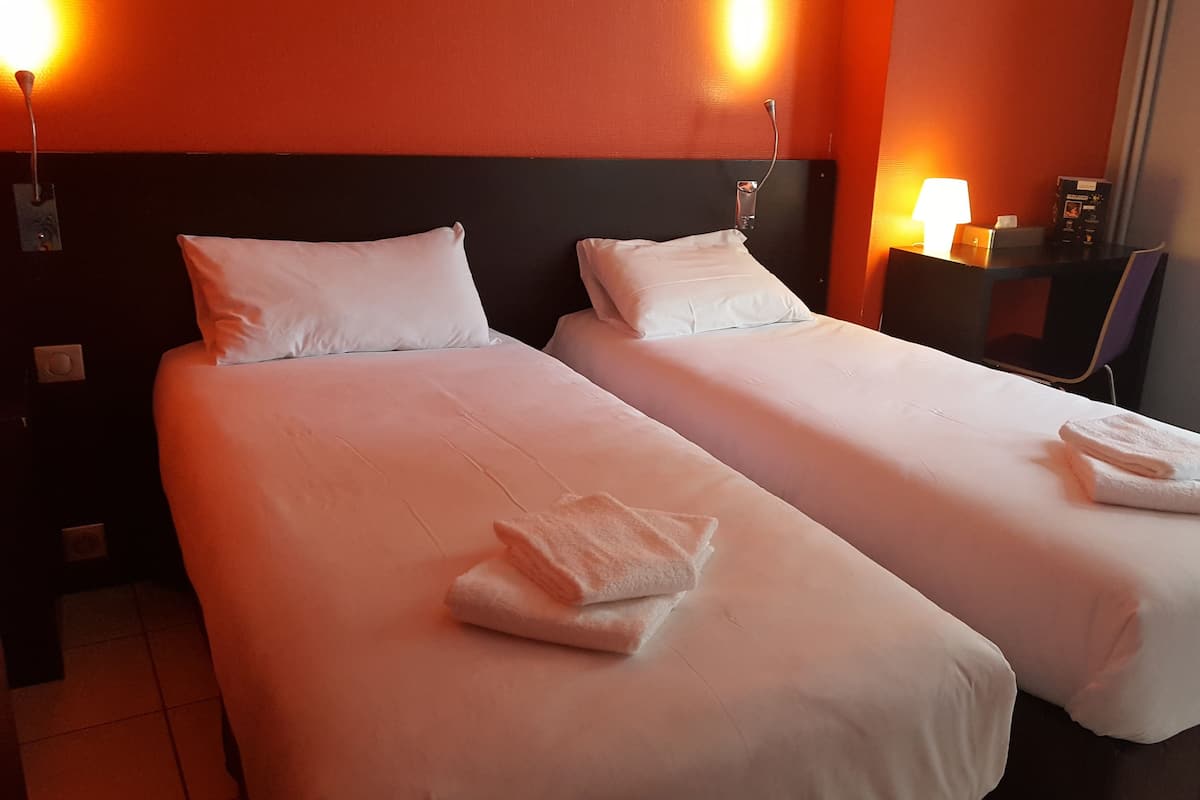 A hotel room with two single beds, each with a white bedspread and pillows, and a small table with a lamp and a box of tissues.