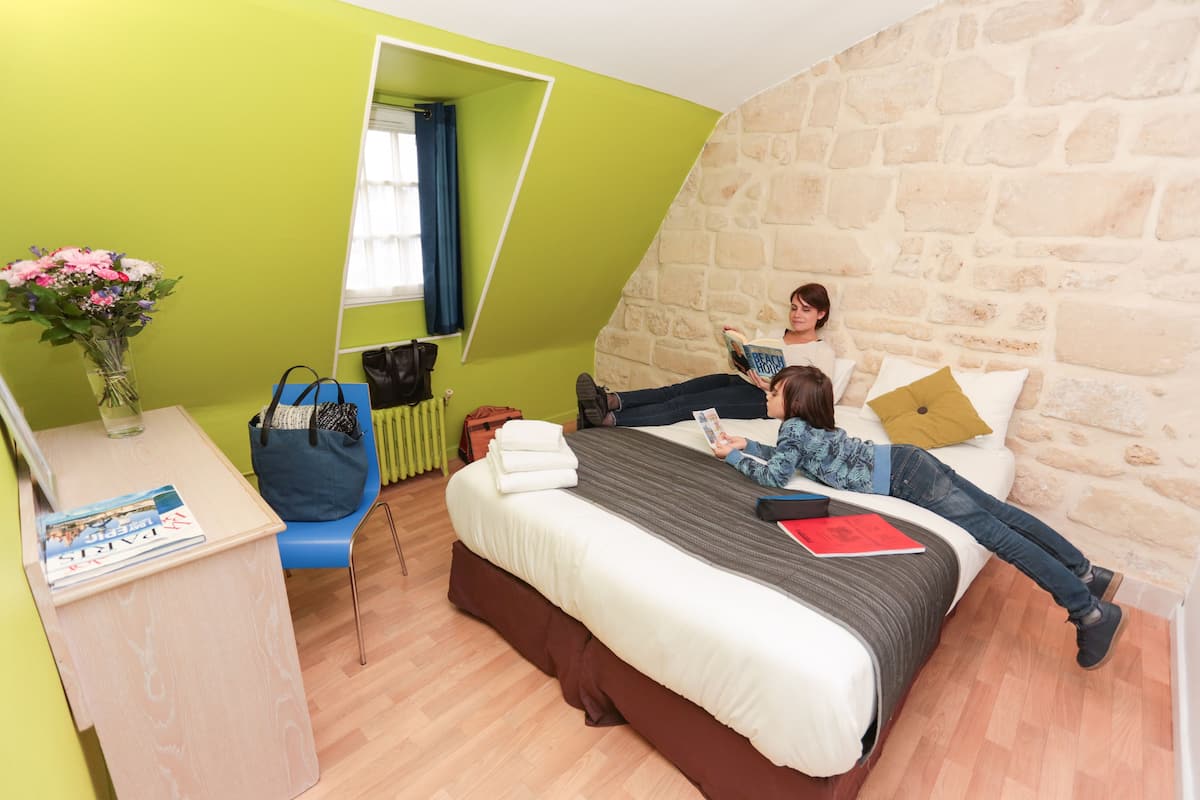 A room with a bed, a desk, a chair, a window with curtains, and a stone wall.