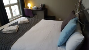 Un lit bien fait, avec du linge de lit blanc et deux serviettes pliées, un bureau avec une lampe et une boîte, une chaise et une fenêtre donnant sur des immeubles.