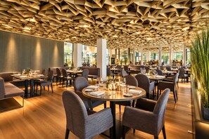 A modern restaurant with a unique geometric ceiling design, wooden flooring, and a mix of round and rectangular tables with chairs. - Grand Hotel Palace Schönbrunn (Vienna)