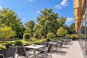 An outdoor patio area with tables and chairs, surrounded by trees and a building with large windows. - Grand Hotel Palace Schönbrunn (Vienna)