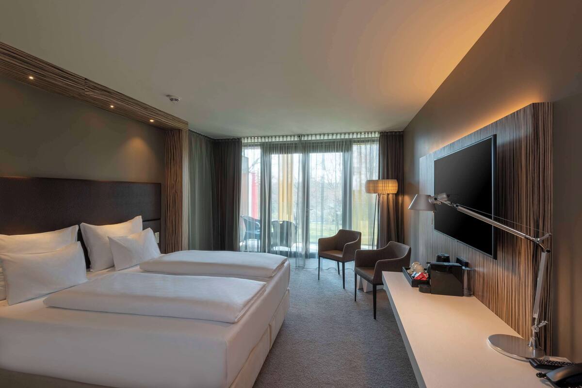 A hotel room with a large bed, a desk, a chair, a TV, and a view of the outdoors through the window.