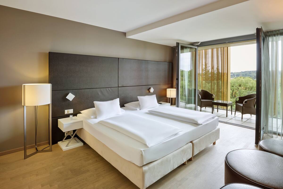 A modern hotel room with a large bed, bedside tables, a lamp, and a balcony with chairs.