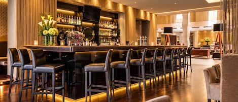 A bar area with a long counter, multiple bar stools, and a variety of bottles.