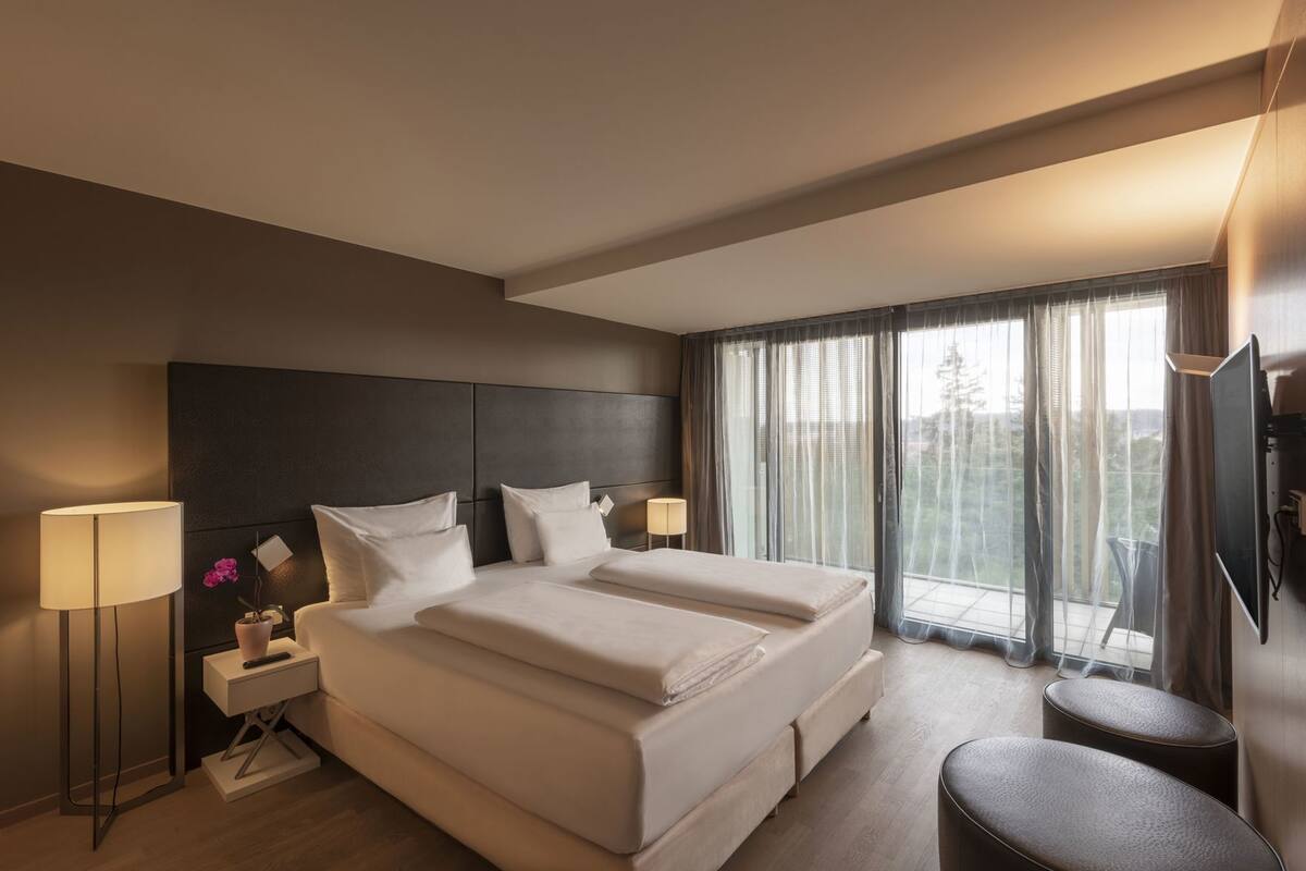 A modern hotel room with a large bed, bedside lamps, a TV, and a balcony with curtains.