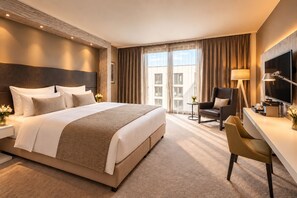 A hotel room with a large bed, a desk, a chair, a TV, and a window with curtains. - Grand Hotel Palace Schönbrunn (Vienna)