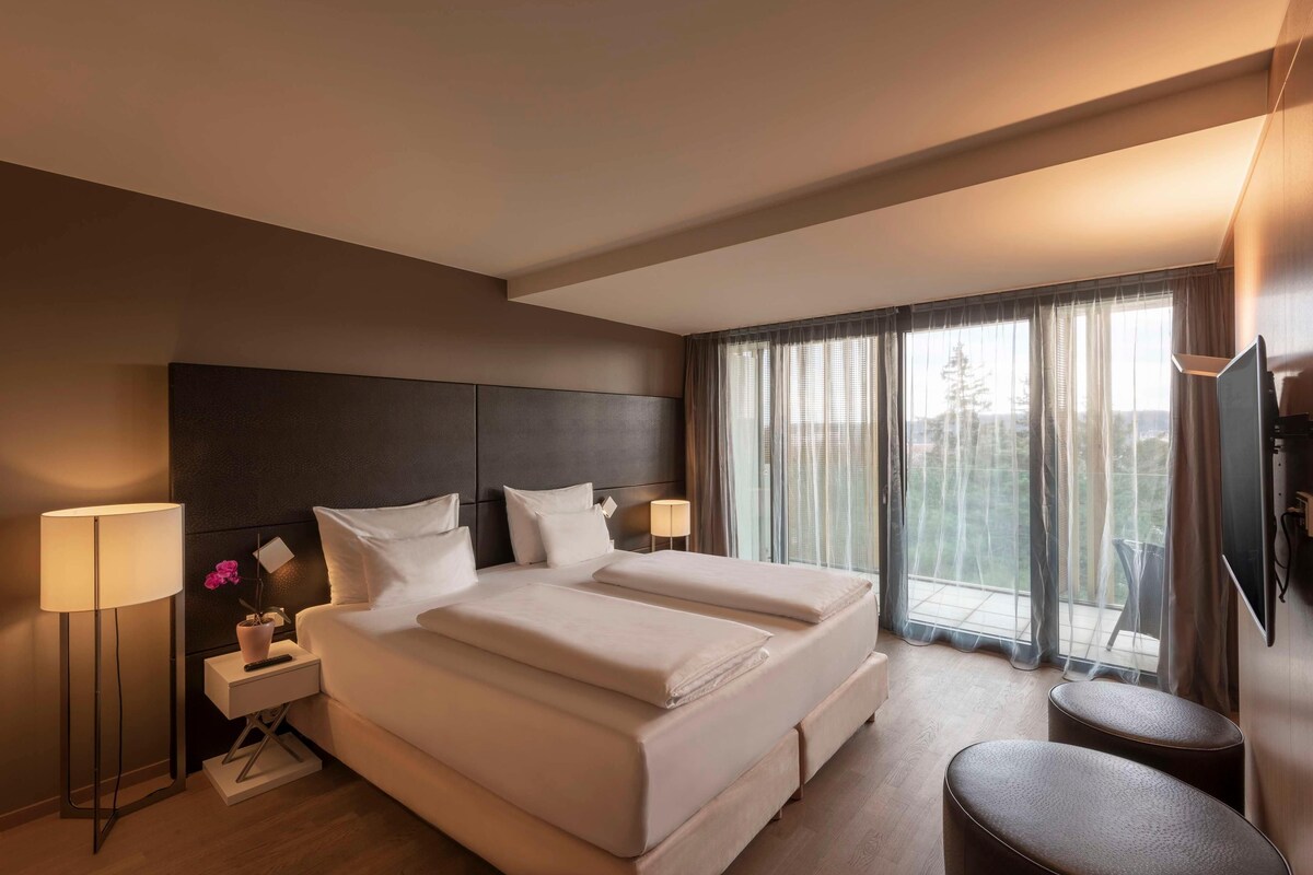 Habitación de hotel con una cama grande, mesitas de noche, televisor y vistas al exterior.