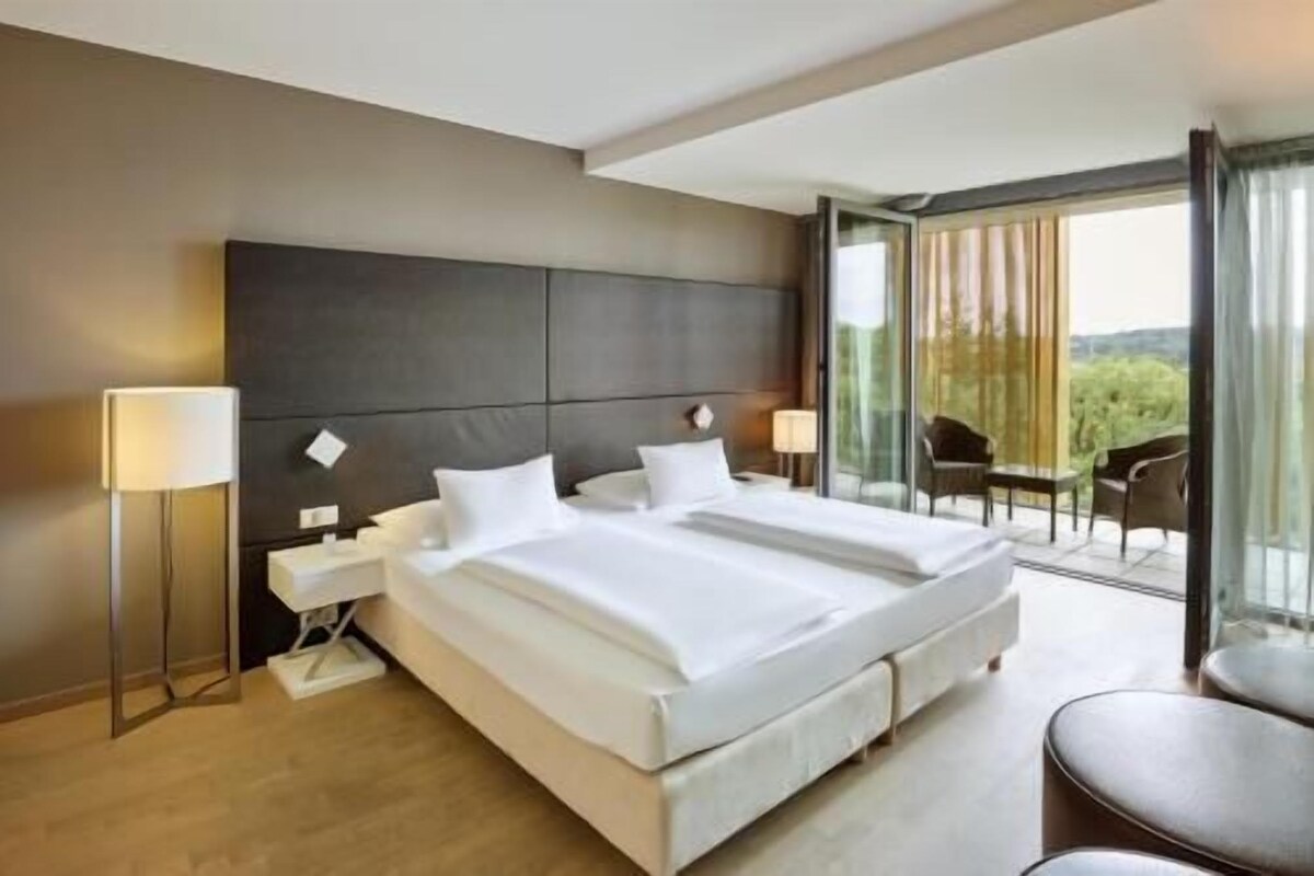 A modern hotel room with a large bed, bedside lamps, a small table, and a balcony with chairs.