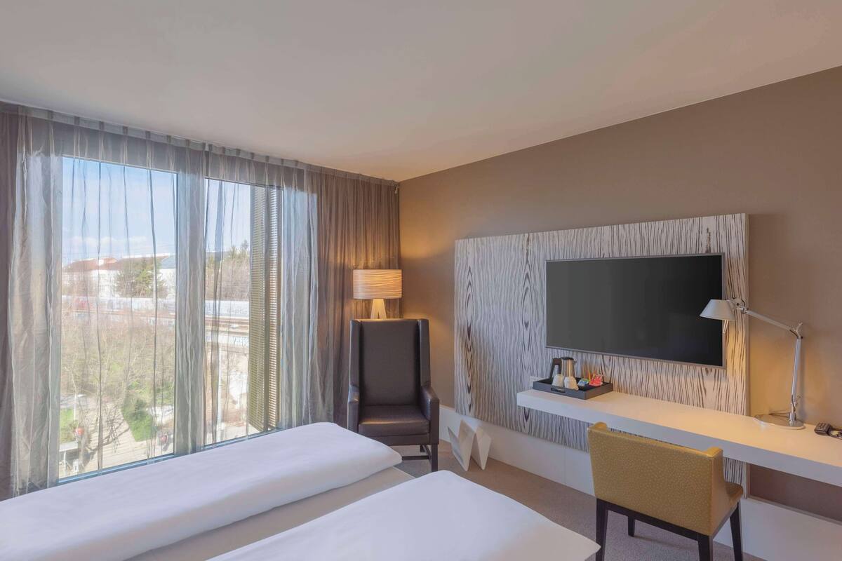 A hotel room with two beds, a desk with a lamp, a TV, and a view of the outside.