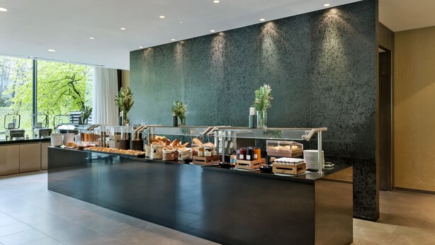 A modern breakfast bar with a variety of food items and beverages, a large window with a view of trees, and a dark textured wall. - Doubletree by Hilton Vienna Schonbrunn (Vienna)