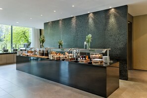 A modern breakfast bar with a variety of food items and beverages, a large window with a view of trees, and a dark textured wall. - Doubletree by Hilton Vienna Schonbrunn (Vienna)