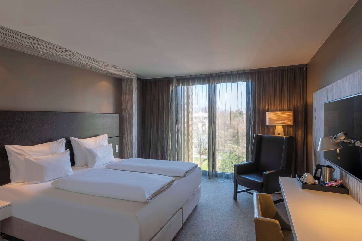 A hotel room with two beds, a desk, a chair, a TV, and a large window with curtains.