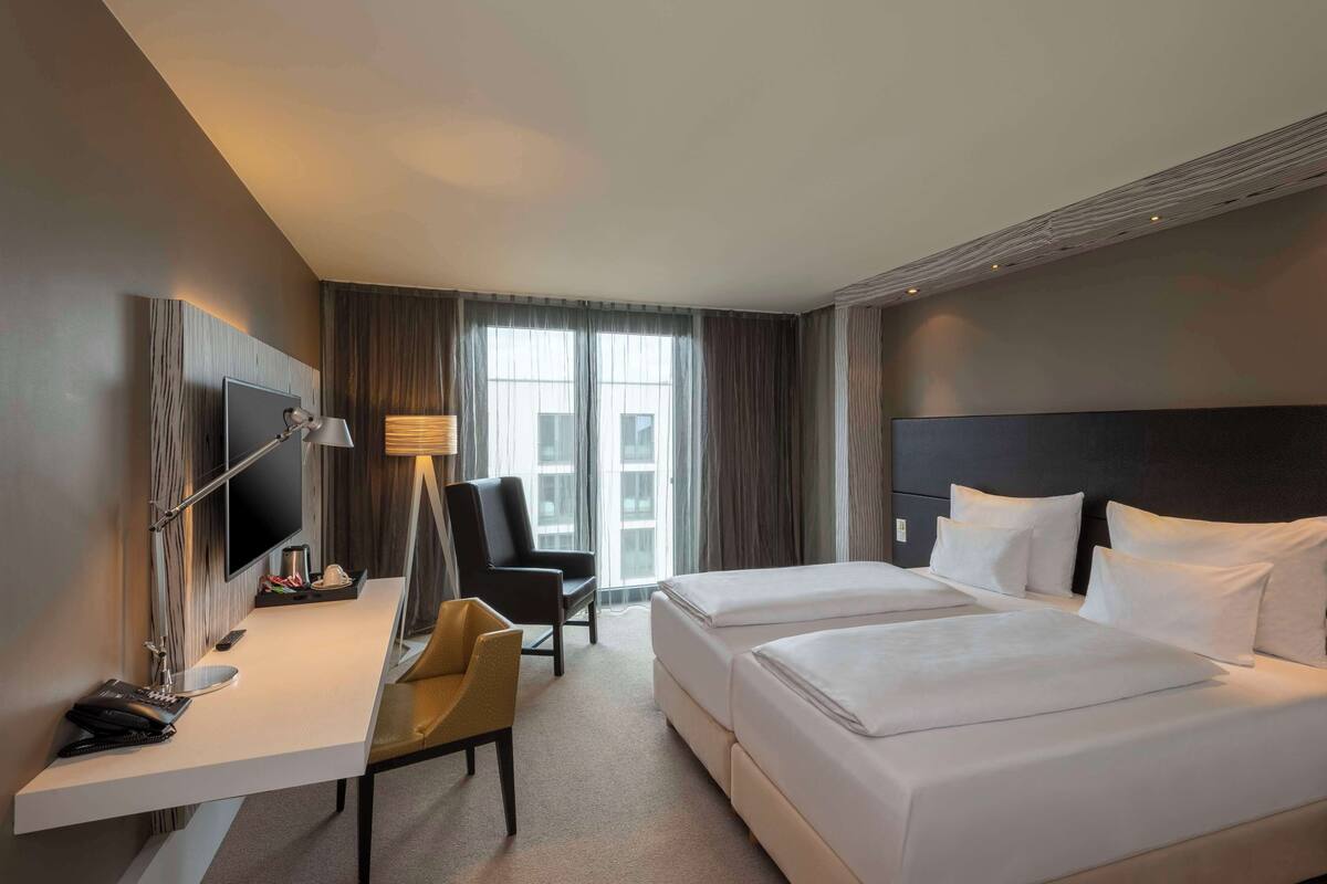 A hotel room with a desk, two beds, a TV, and a large window with curtains.
