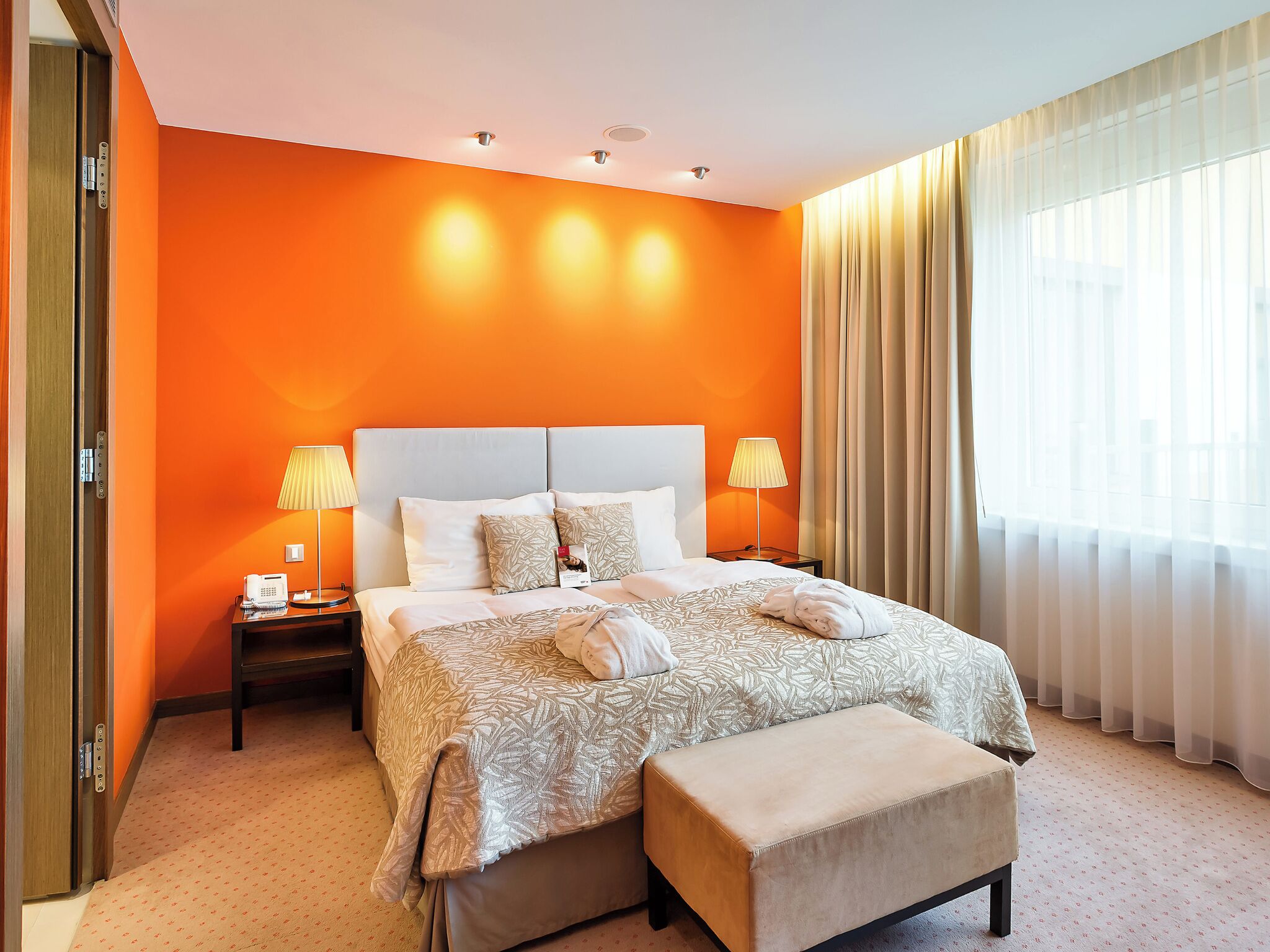 Een hotelkamer met een groot bed, twee bedlampen, een bedtelefoon, een raam met dunne gordijnen en een deur met lift.