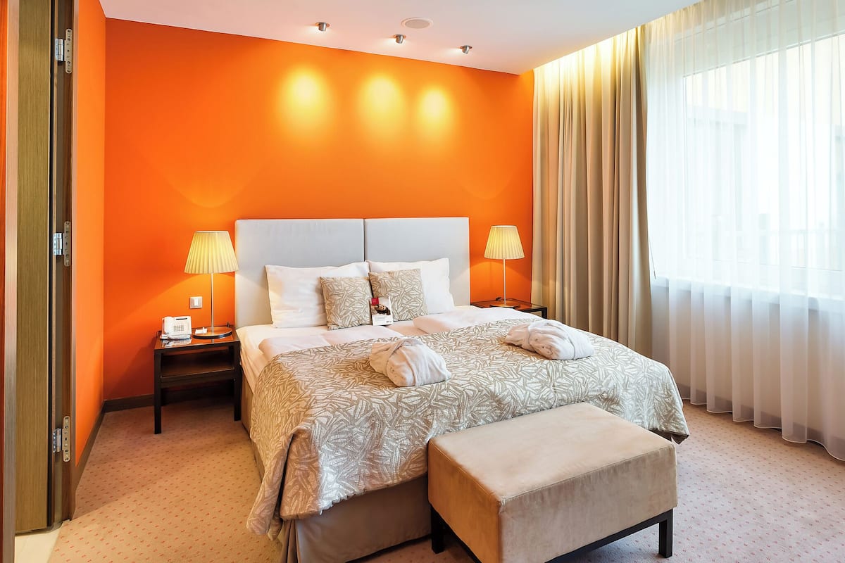 Ein Hotelzimmer mit einem großen Bett, zwei Nachttischlampen, einem Telefon am Nachttisch, einem Fenster mit durchsichtigen Vorhängen und einer Tür mit Aufzug.