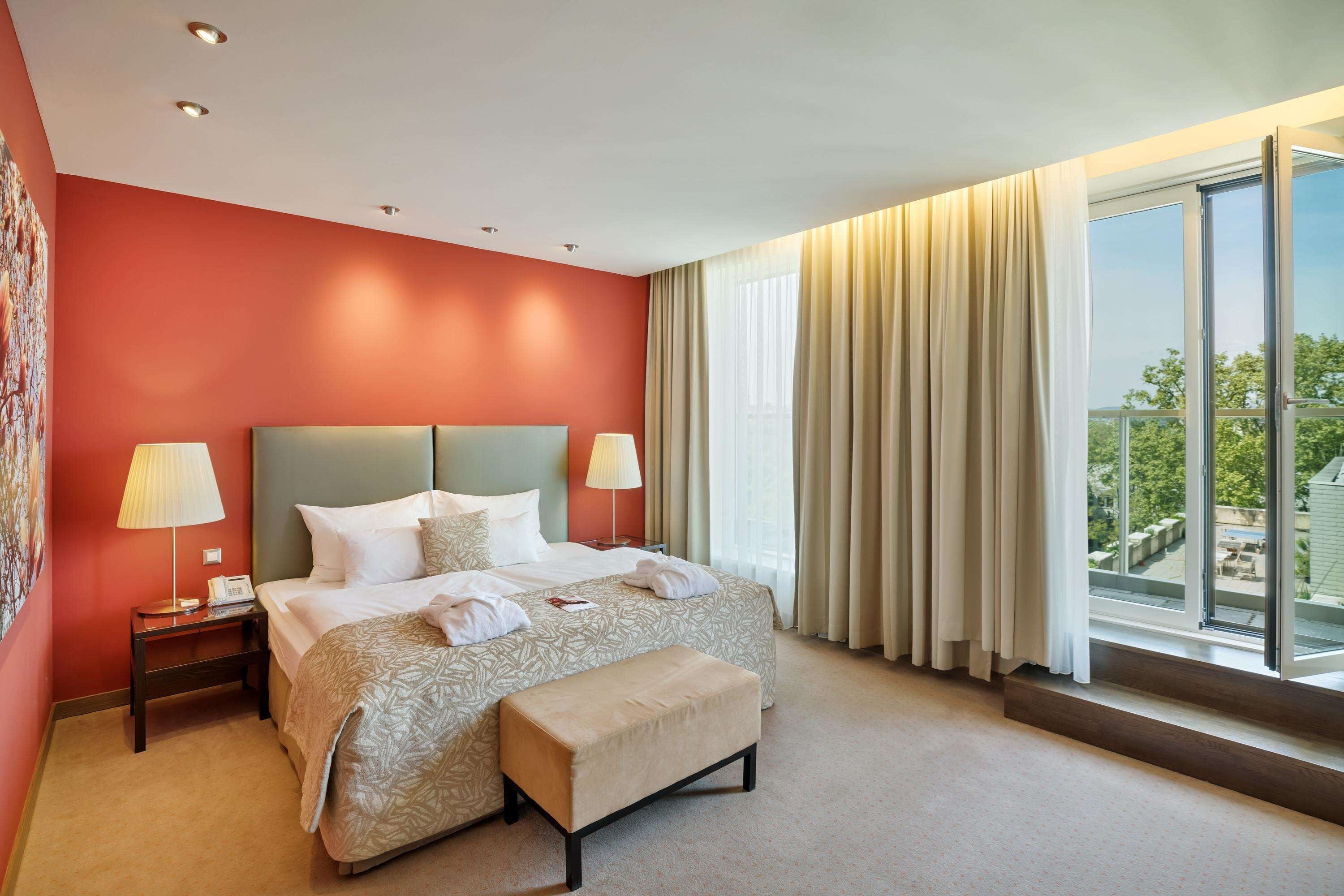 Quarto de hotel com uma cama grande, duas mesas de cabeceira com abajures, um banco e uma janela ampla com cortinas.