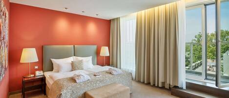 Quarto de hotel com uma cama grande, duas mesas de cabeceira com abajures, um banco e uma janela ampla com cortinas.