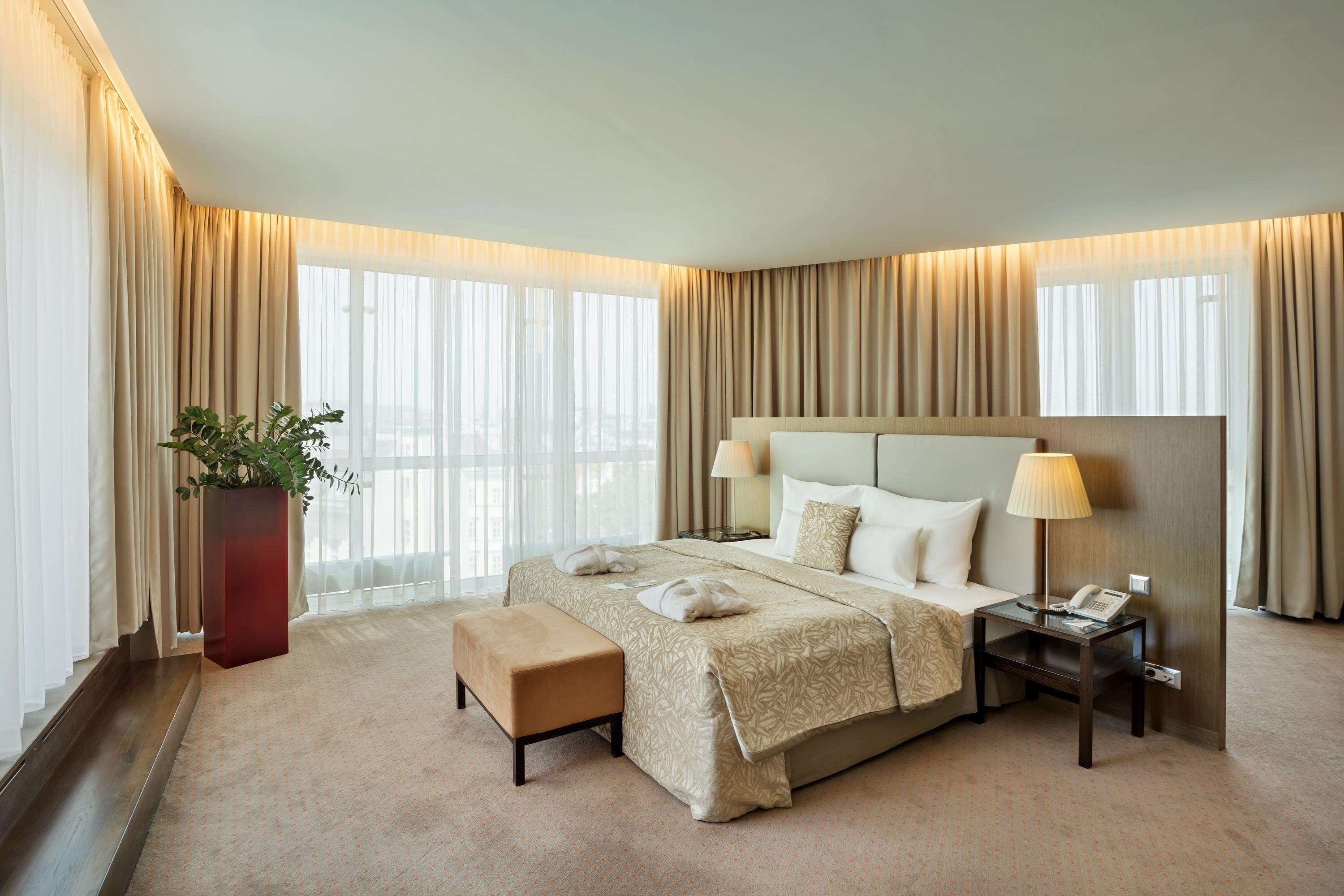 Quarto de hotel com uma cama grande, mesa de cabeceira com abajur, pequena mesa com telefone, planta em vaso e vista da cidade por grandes janelas.