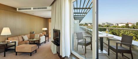 Una habitación de hotel moderna con sofá, sillones, televisor y balcón con mesa y sillas.
