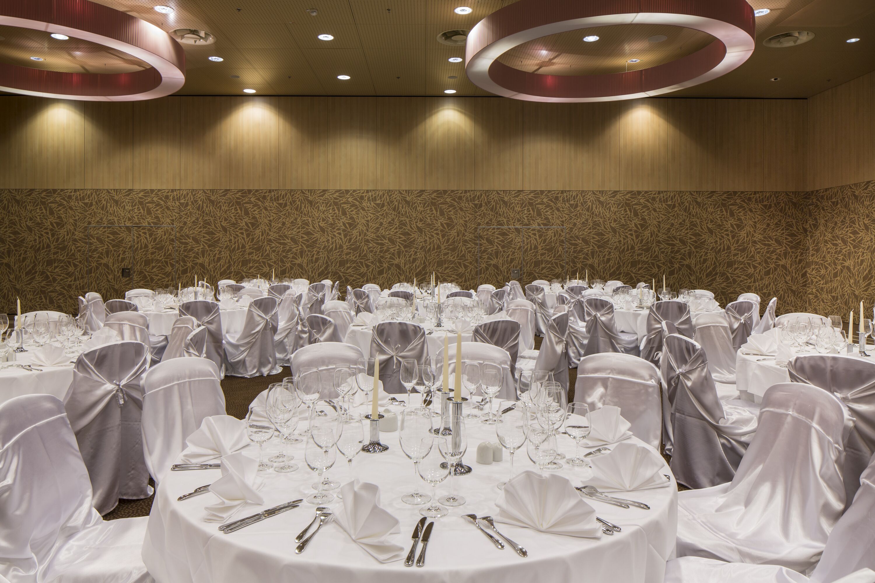 A banquet hall with round tables set for a formal event, featuring white tablecloths, silverware, and candle holders.