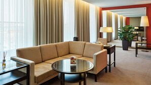 A modern hotel room with a beige sofa, a glass coffee table, and a dining area with a lamp and potted plant.