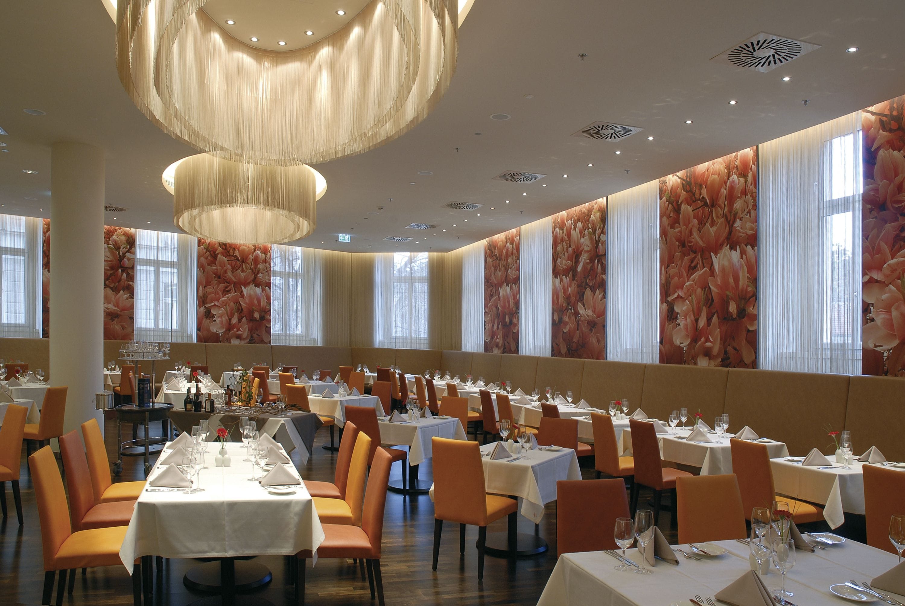 Uma ampla sala de jantar com várias mesas e cadeiras, janelas grandes com cortinas florais e um lustre moderno.