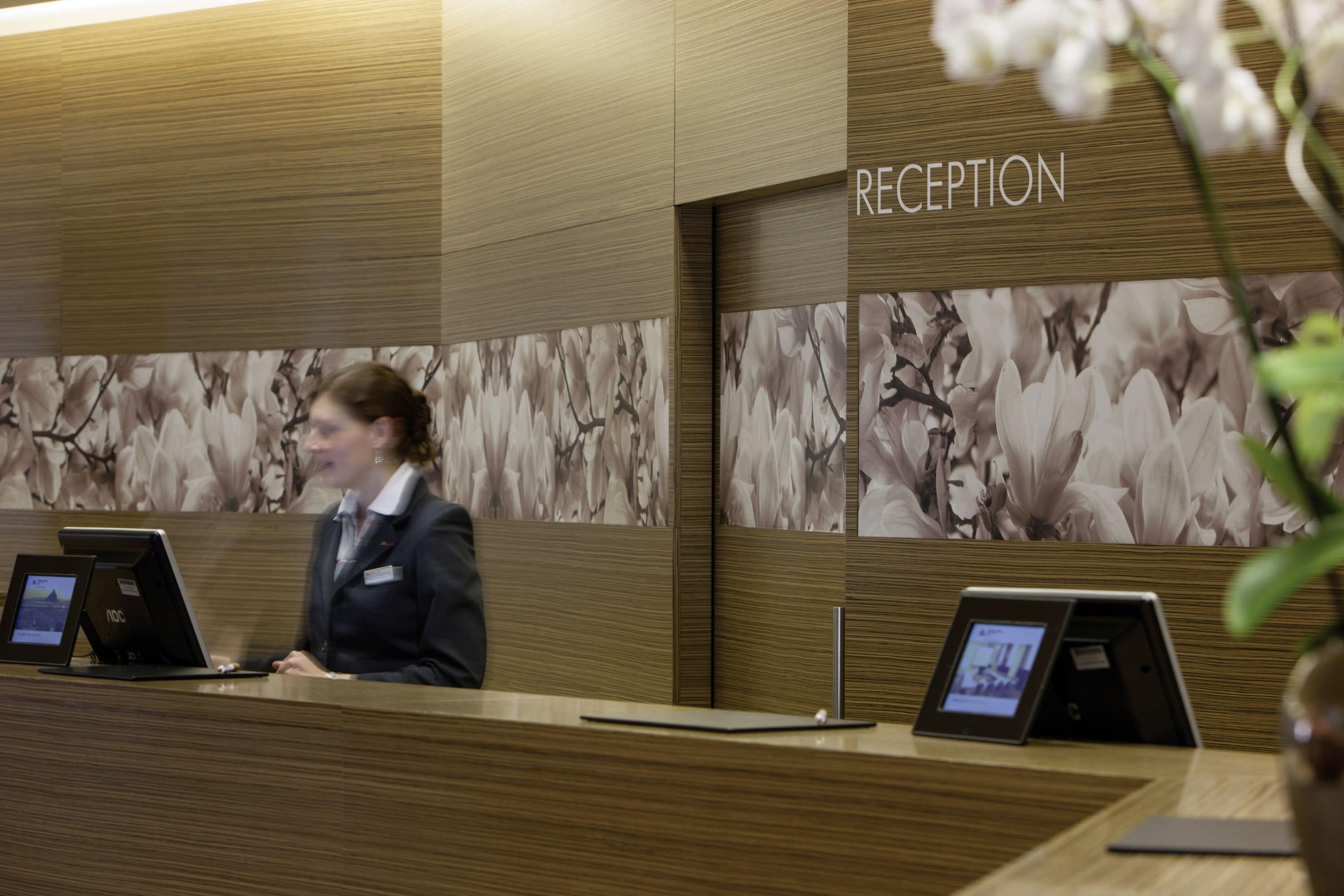 A reception area with a wooden reception desk, a digital display, and a floral wallpaper backdrop.