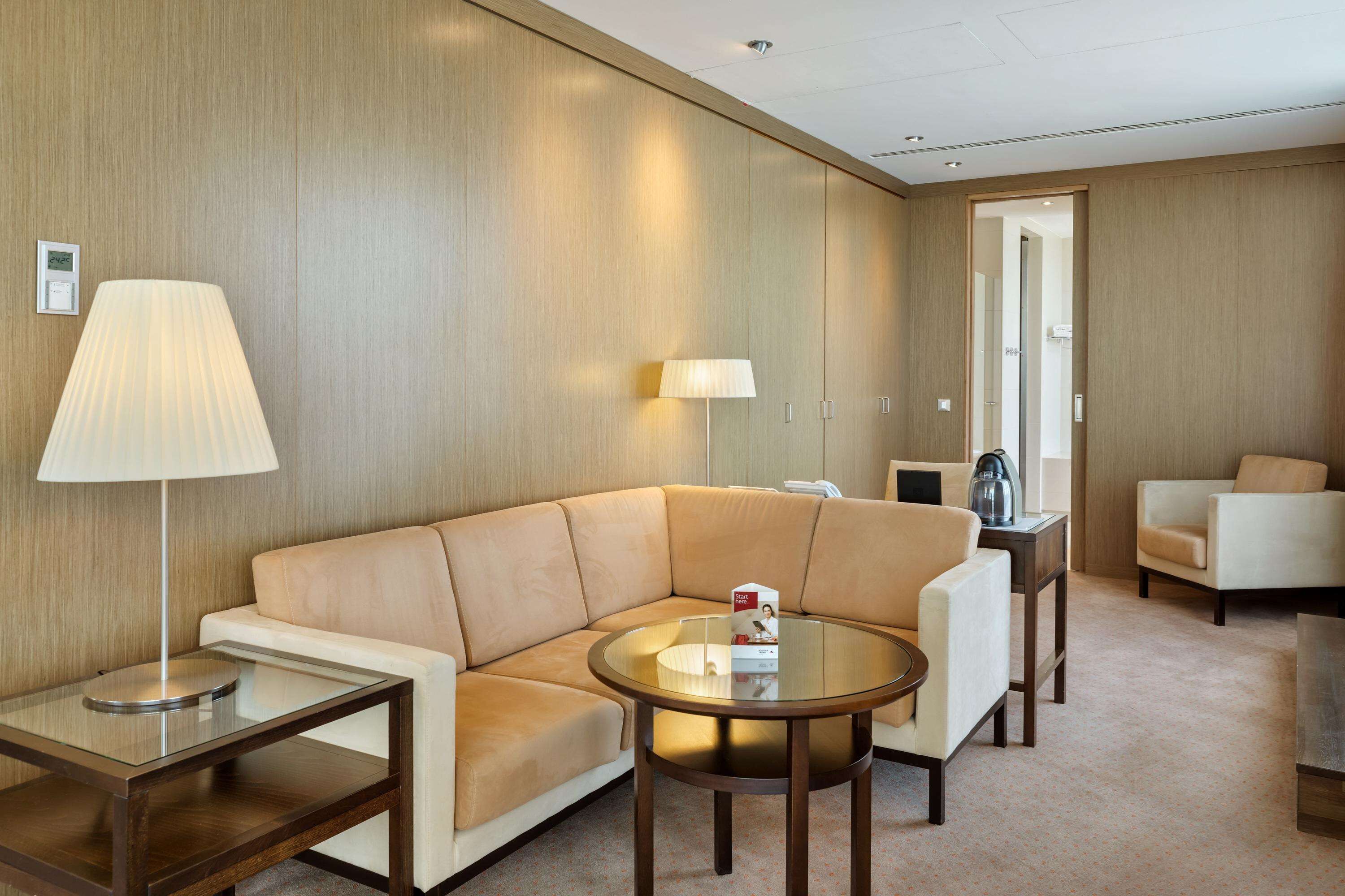 A modern hotel room with a beige leather sofa, a glass coffee table, a lamp, and a side table.