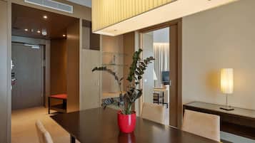 A modern hotel room with a wooden dining table, a red vase with a plant, a glass shelf, and a desk with a lamp.