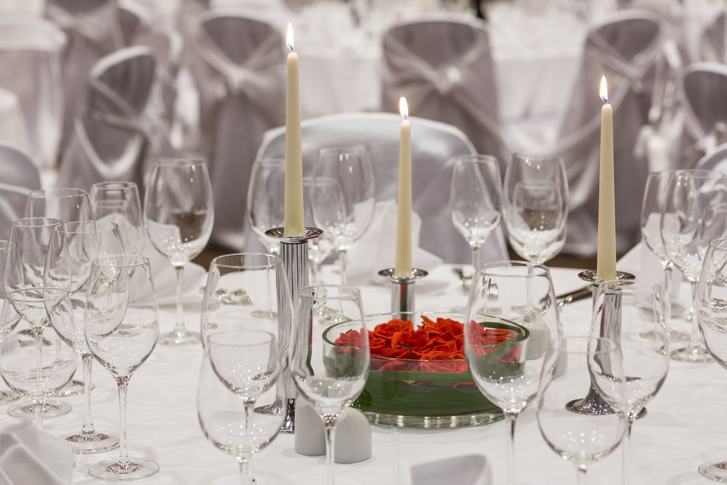 Ett bord dukat för ett formellt evenemang, med klara glas, vita ljus och en mittdekoration bestående av röda blommor.