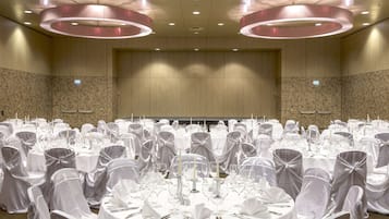 Uma sala de banquetes com mesas redondas preparadas para um evento formal, com toalhas brancas, talheres e copos.