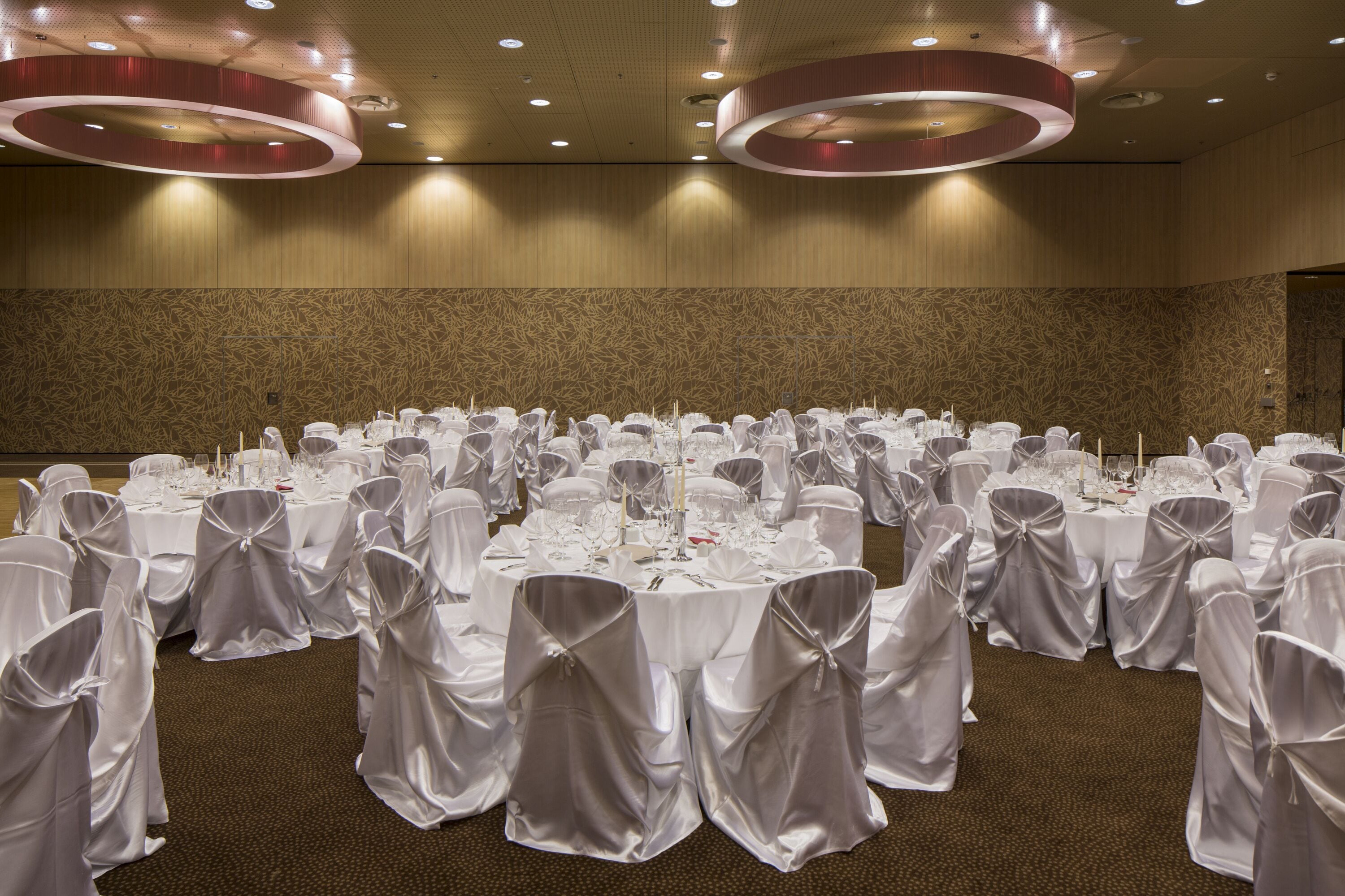 Salão de festas com mesas redondas preparadas para um evento formal, com toalhas brancas e cadeiras adornadas com fitas brancas.