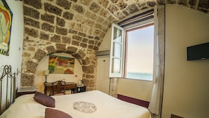 A bedroom with a stone wall, a bed with a headboard, a desk, a chair, a television, and a window with a view of the sea. - Charme Hotel Henry's House (Syracuse)
