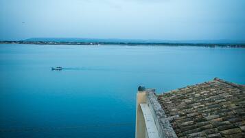 Una masa de agua tranquila con un barco a lo lejos, el borde de un tejado de baldosas y una orilla distante con edificios.