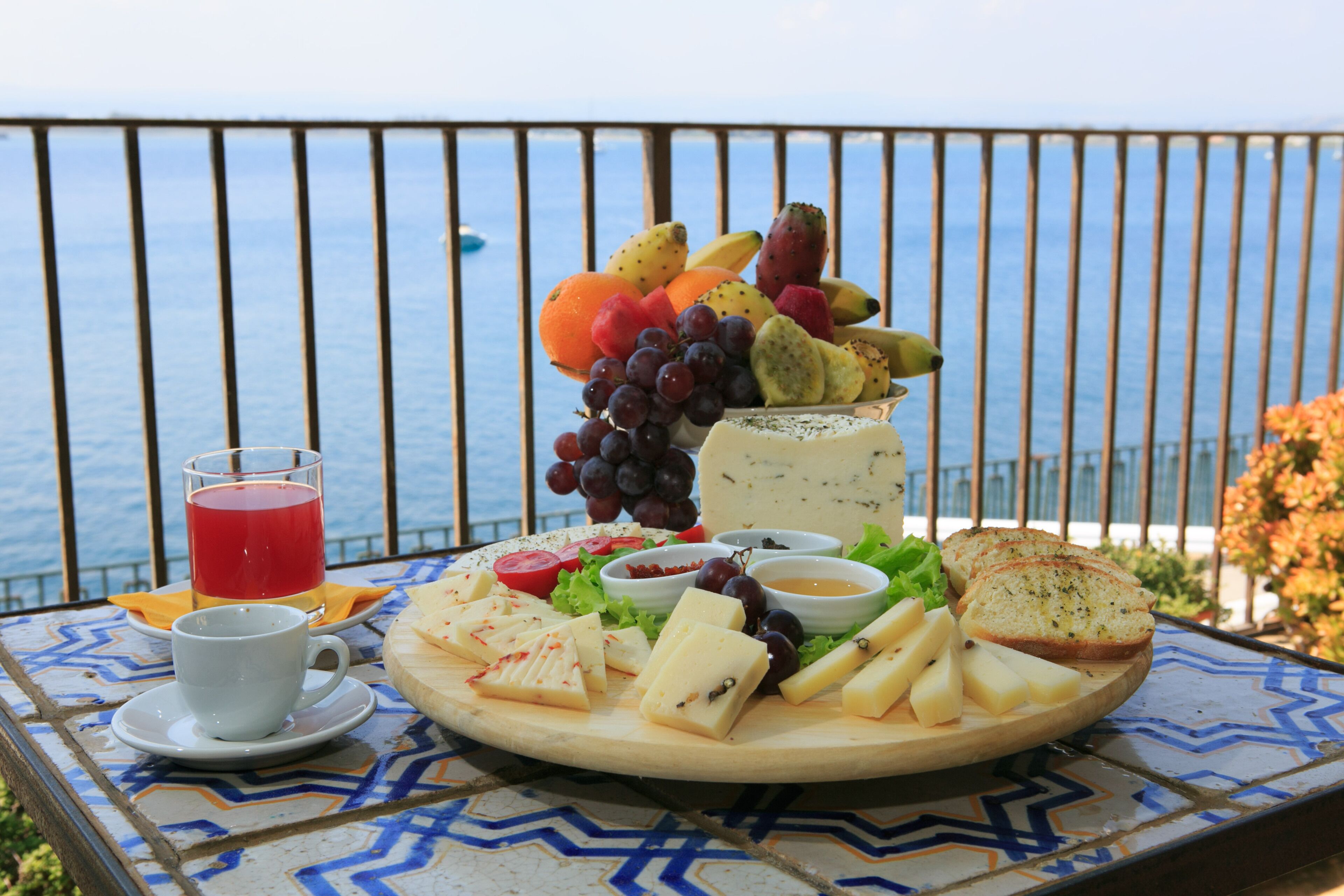 A table with a cheese board, fruits, and a glass of red drink, set against a balcony with a view of the sea.