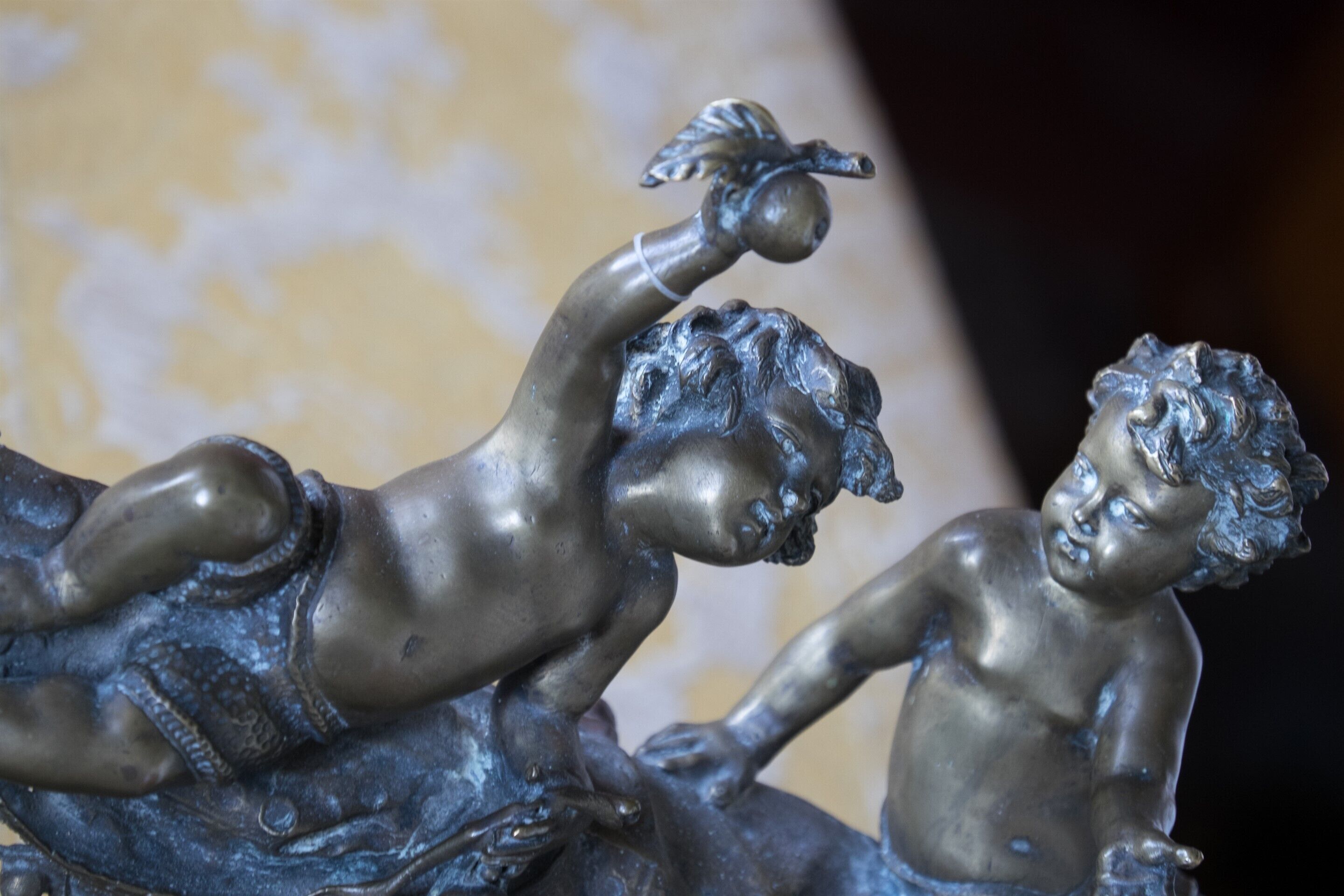 Une sculpture en bronze représentant deux figures : l’une est allongée et l’autre est debout, avec une feuille et des fruits en arrière-plan.
