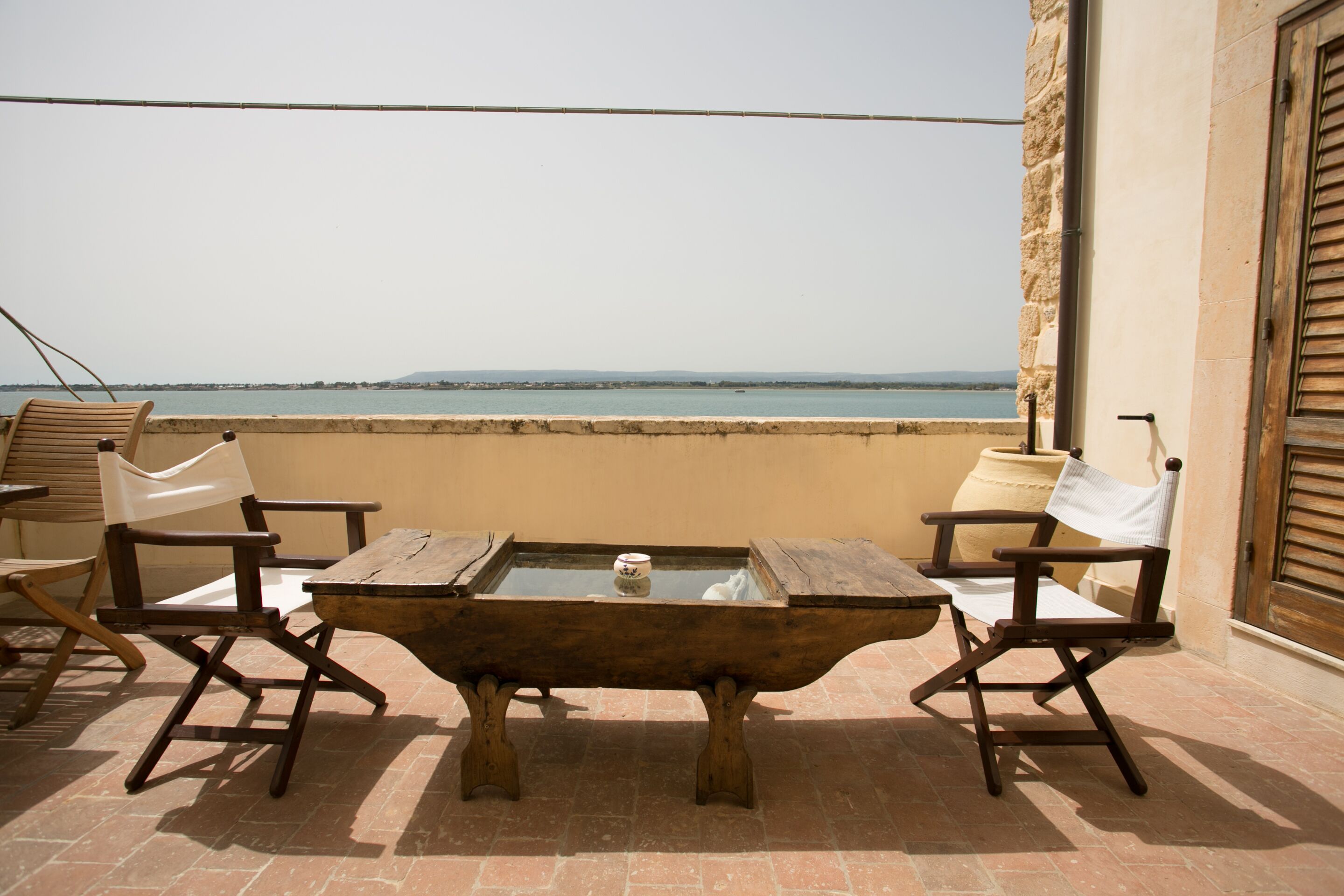 A terrace with a wooden table and chairs, a view of the sea, and a stone wall.