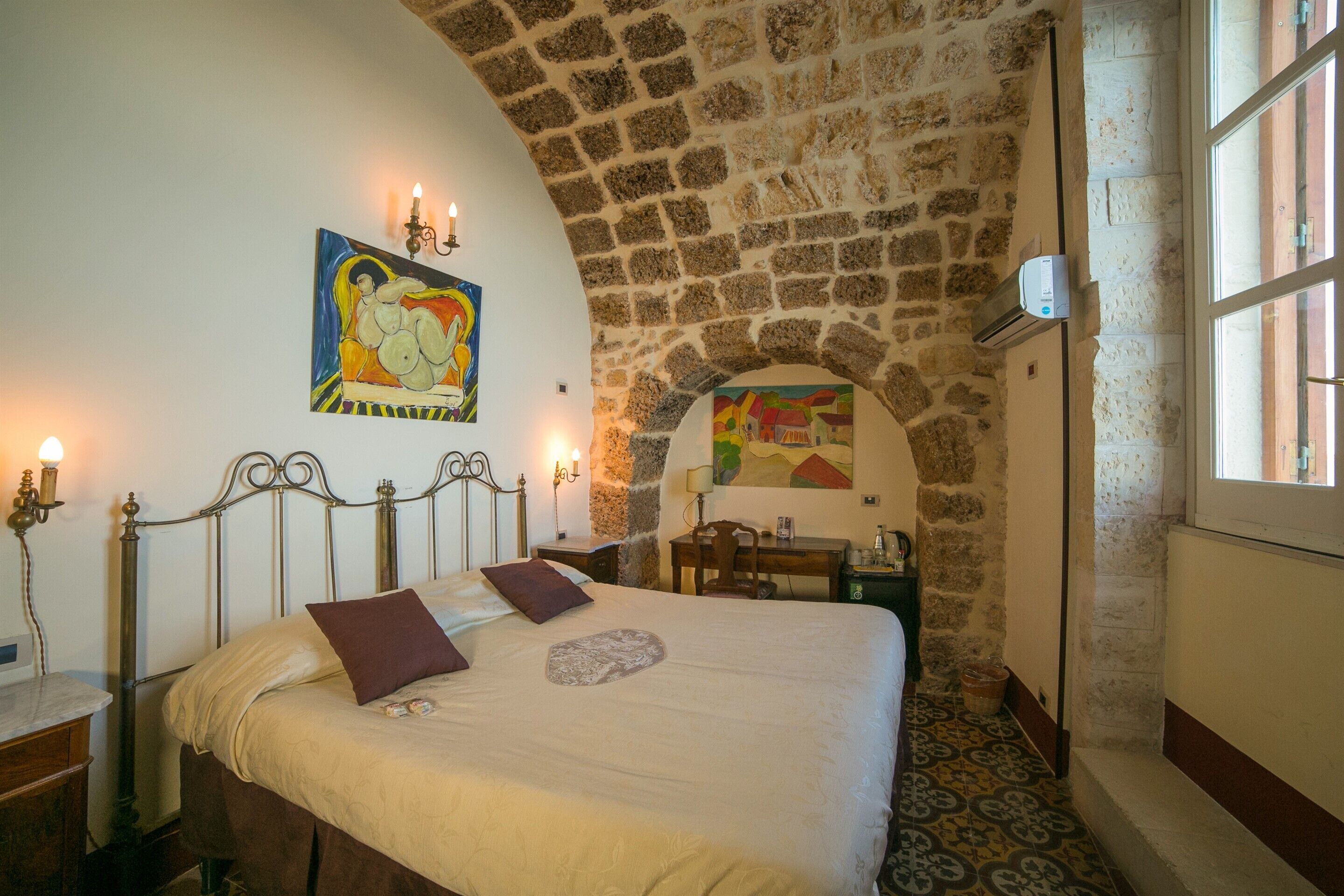 A bedroom with a stone wall, a bed with a headboard, a desk, and a painting on the wall.