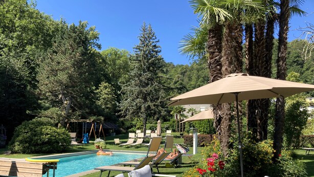 A pool area with lounge chairs and umbrellas, surrounded by greenery and trees. - Villa Siesta, Park & Pool Living (Losone)