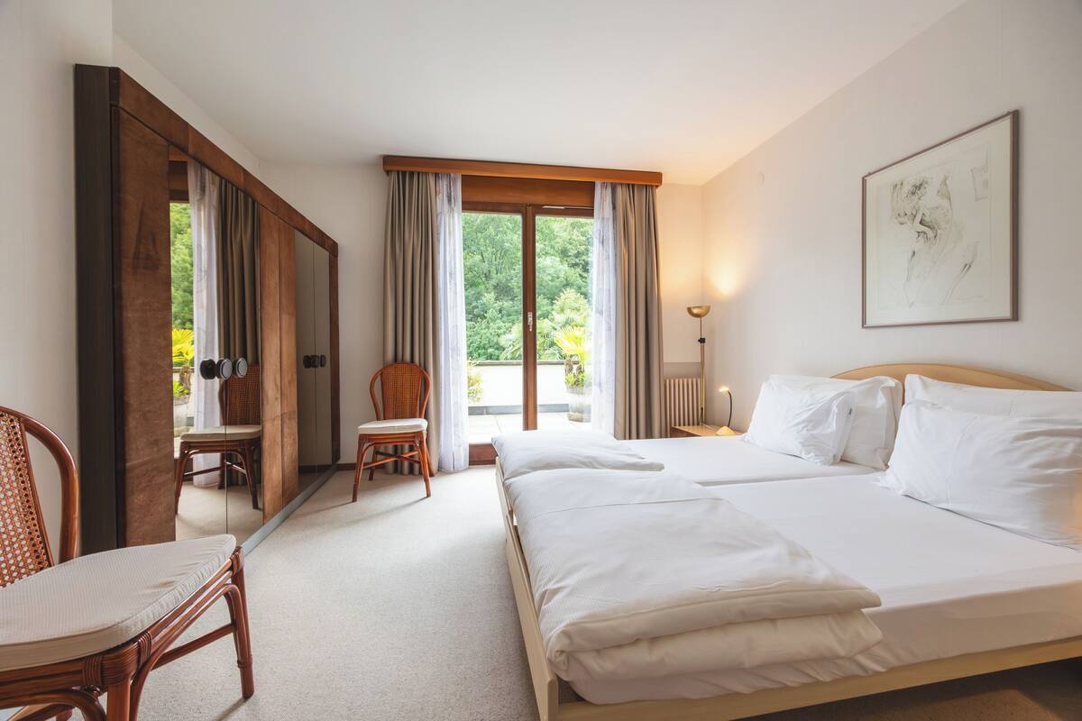 Quarto de hotel com uma cama grande, uma cadeira, uma mesa e vista para o verde pela janela.