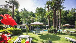 A garden with palm trees, a pool, lounge chairs, and a small building. - Villa Siesta, Park & Pool Living (Losone)
