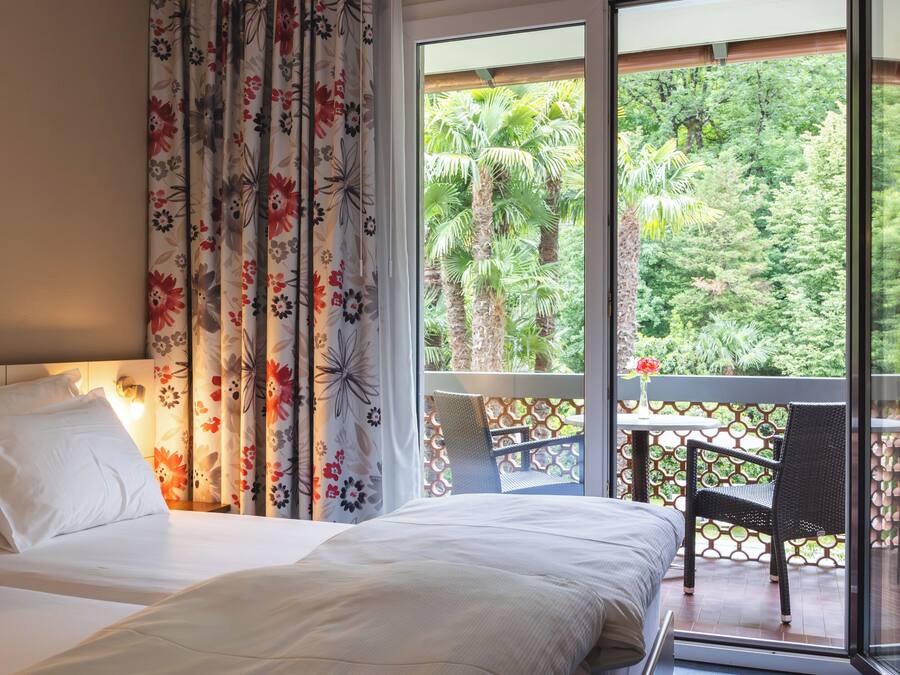 Une chambre d’hôtel avec un lit, une chaise, une fenêtre donnant sur un paysage verdoyant et un rideau orné d’un motif floral.